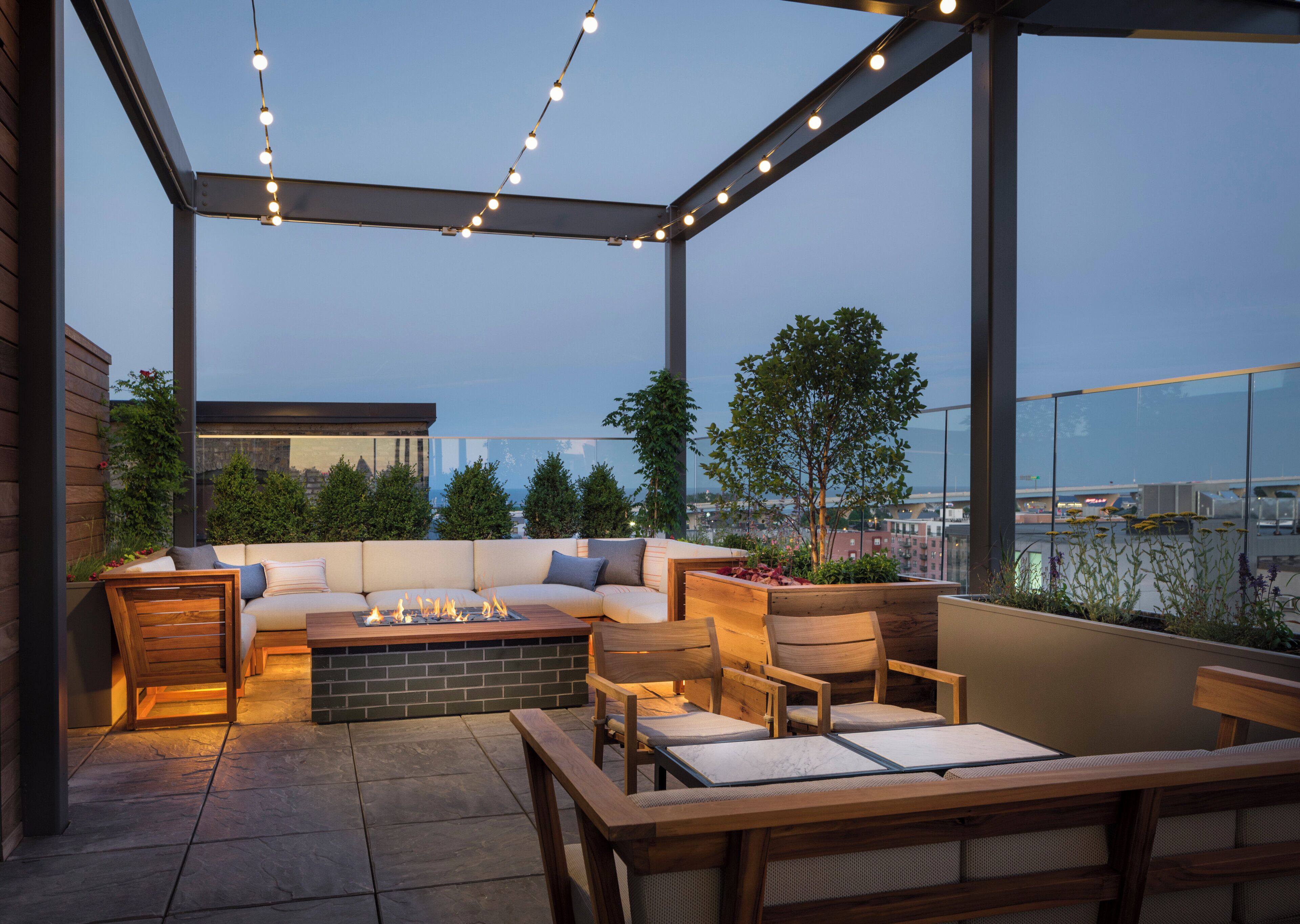 A rooftop patio with a sofa set, wooden chairs, a fire pit, and string lights.