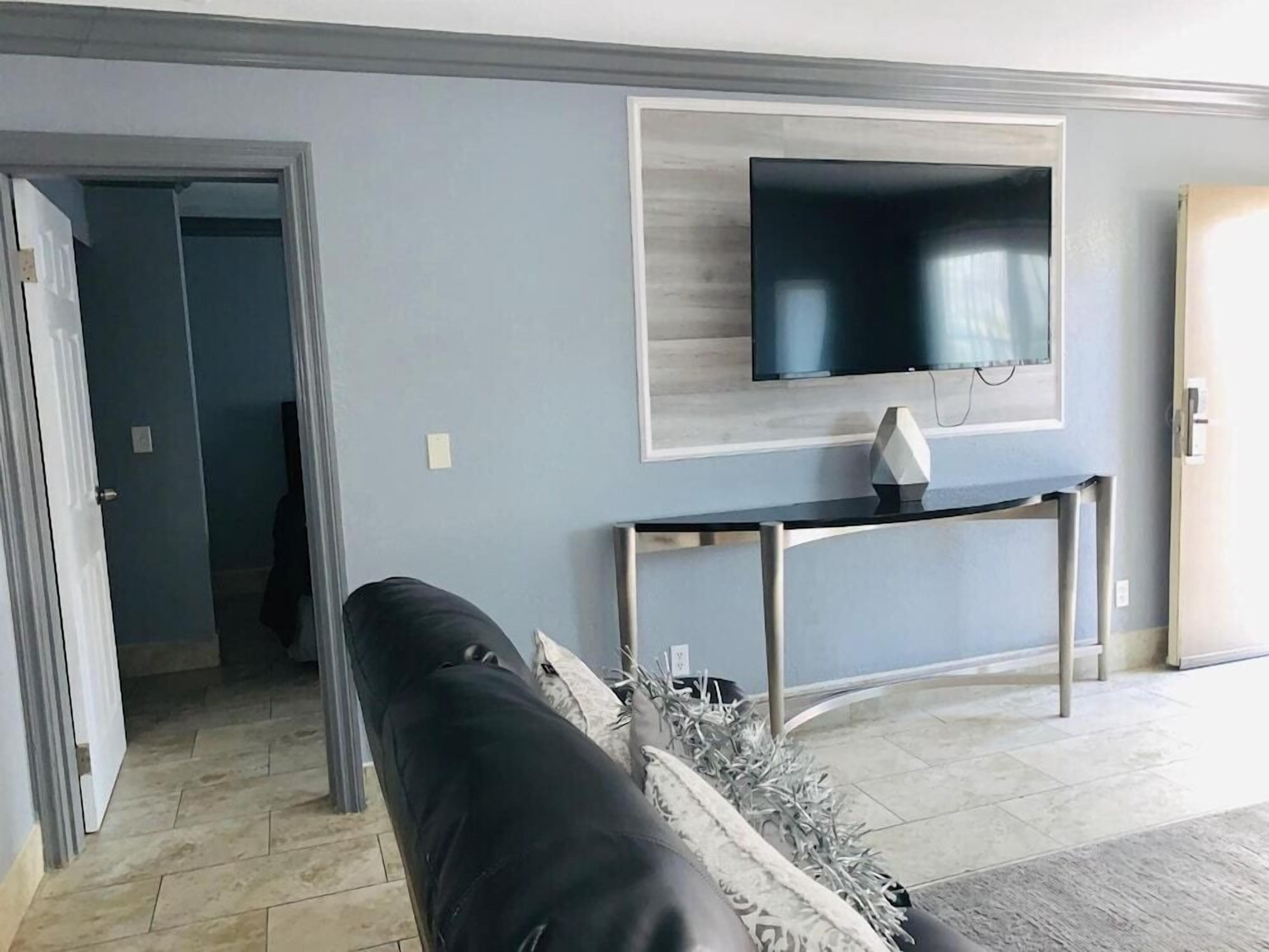 Uma sala de estar com um sofá preto, uma TV fixada na parede e uma pequena mesa.