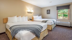 A hotel room with two beds, a nightstand, a wall-mounted lamp, a window with a view of greenery, and a radiator.