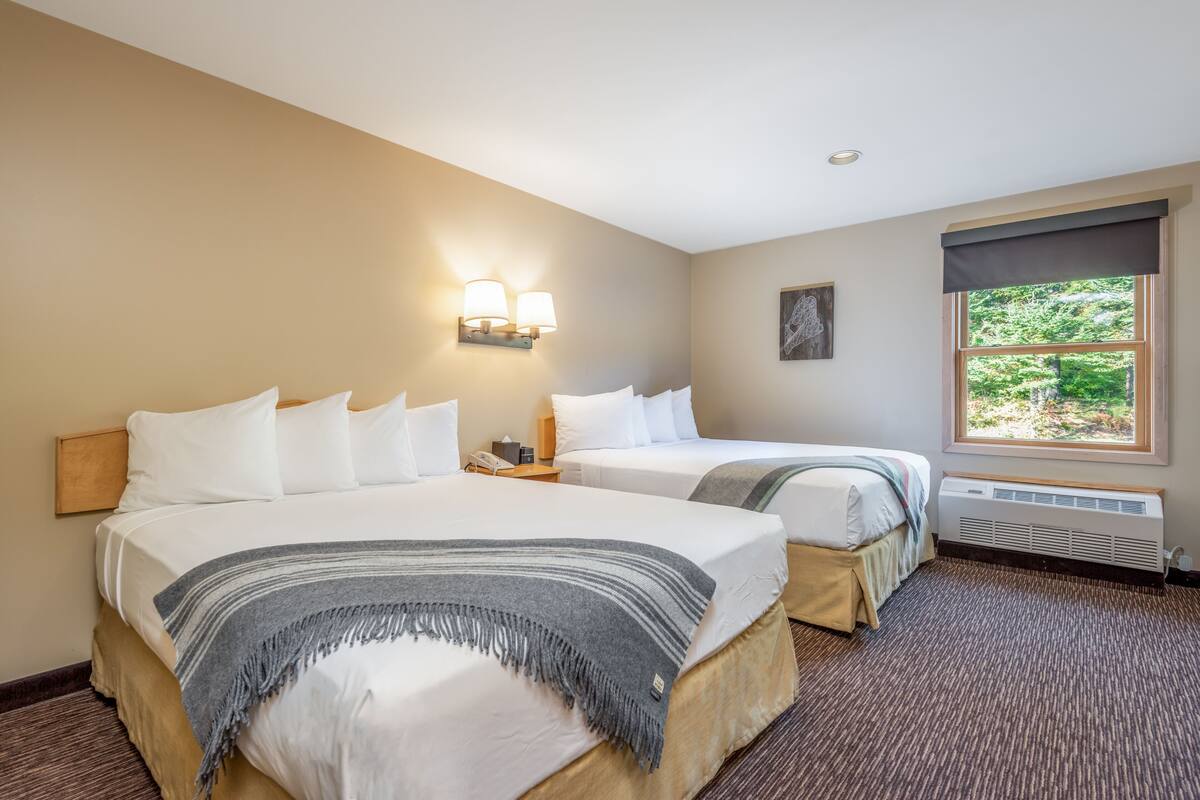 A hotel room with two beds, a nightstand, a wall-mounted lamp, a window with a view of greenery, and a radiator.