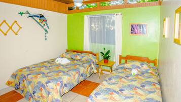 A room with two single beds, a small table with a plant, a ceiling fan, and a window with curtains.