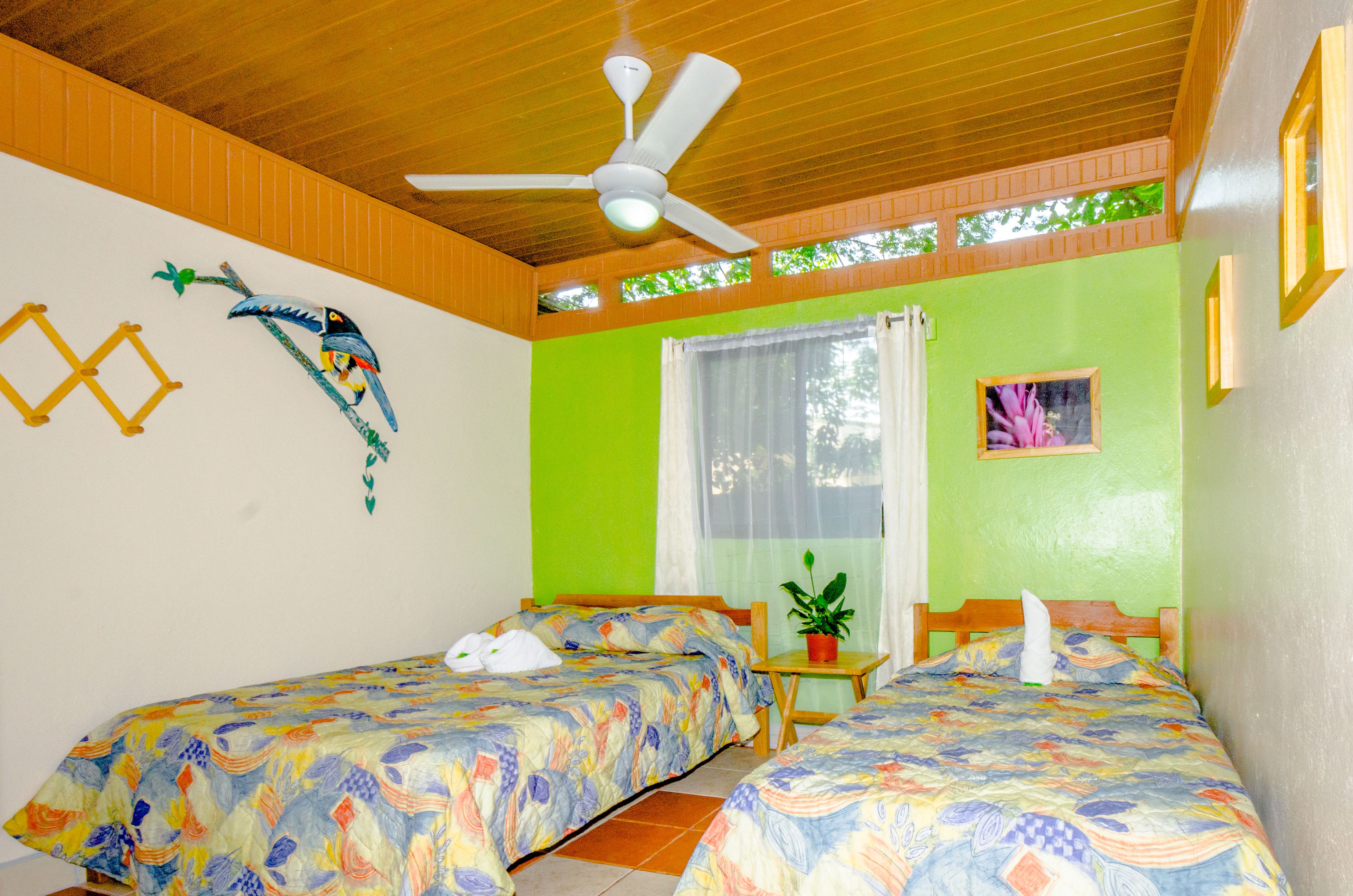 Una habitación con dos camas, un ventilador de techo, una ventana con cortinas, una mesita con una planta y un adorno en la pared.