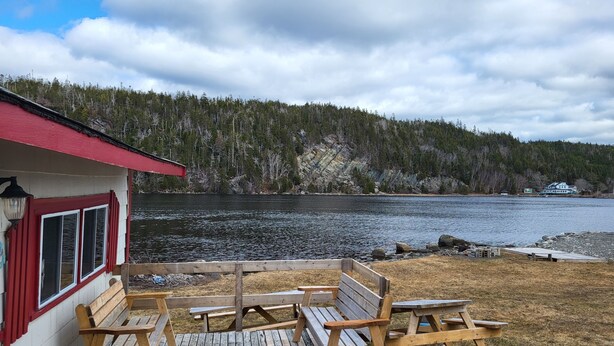 A wooden deck with benches overlooking a lake, a red building with windows, and a forested hillside in the background. - Jeddore Lodge Cabins (Head of Jeddore)