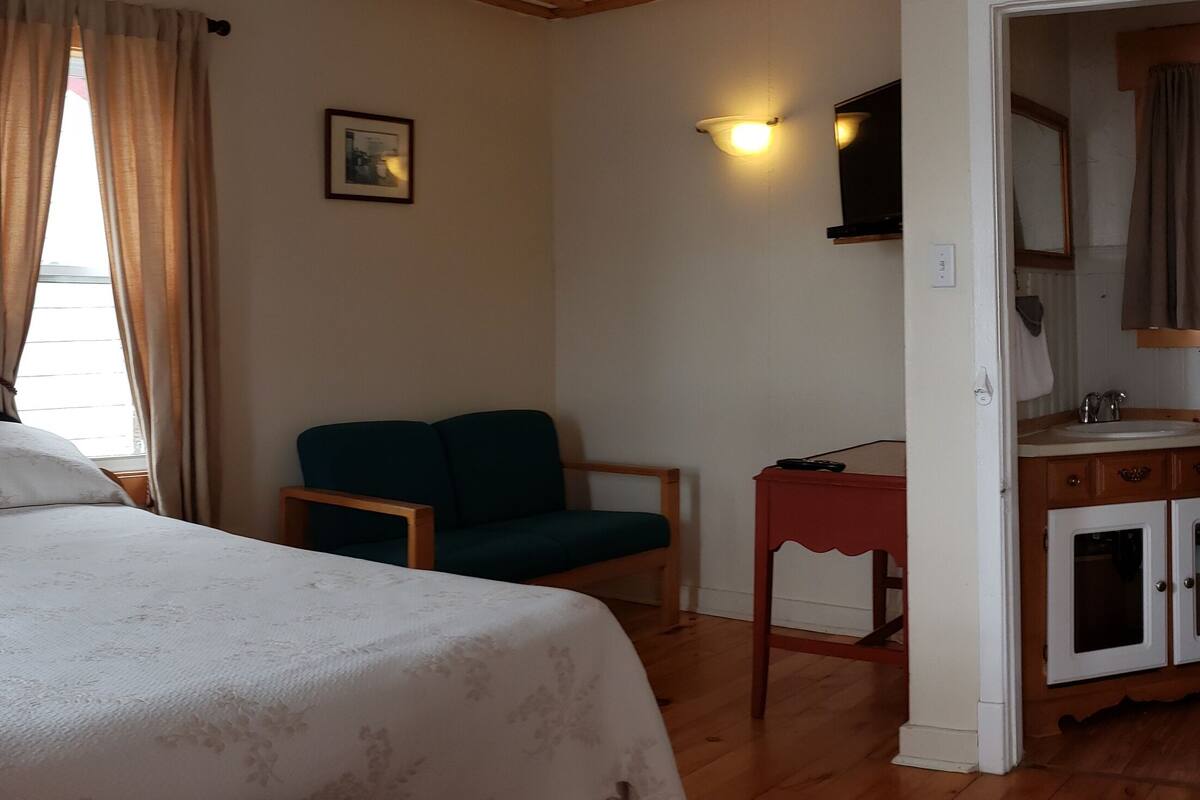 Ein Schlafzimmer mit Bett, Sessel, Nachttisch, Lampe, einem Fenster mit Vorhängen und einem Badezimmer mit Waschbecken und Spiegel.