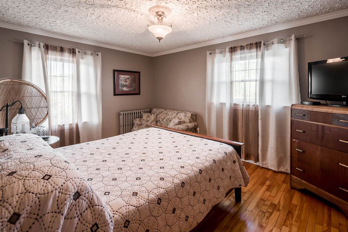 A bedroom with a bed, a dresser, a television, a radiator, and a window with curtains.