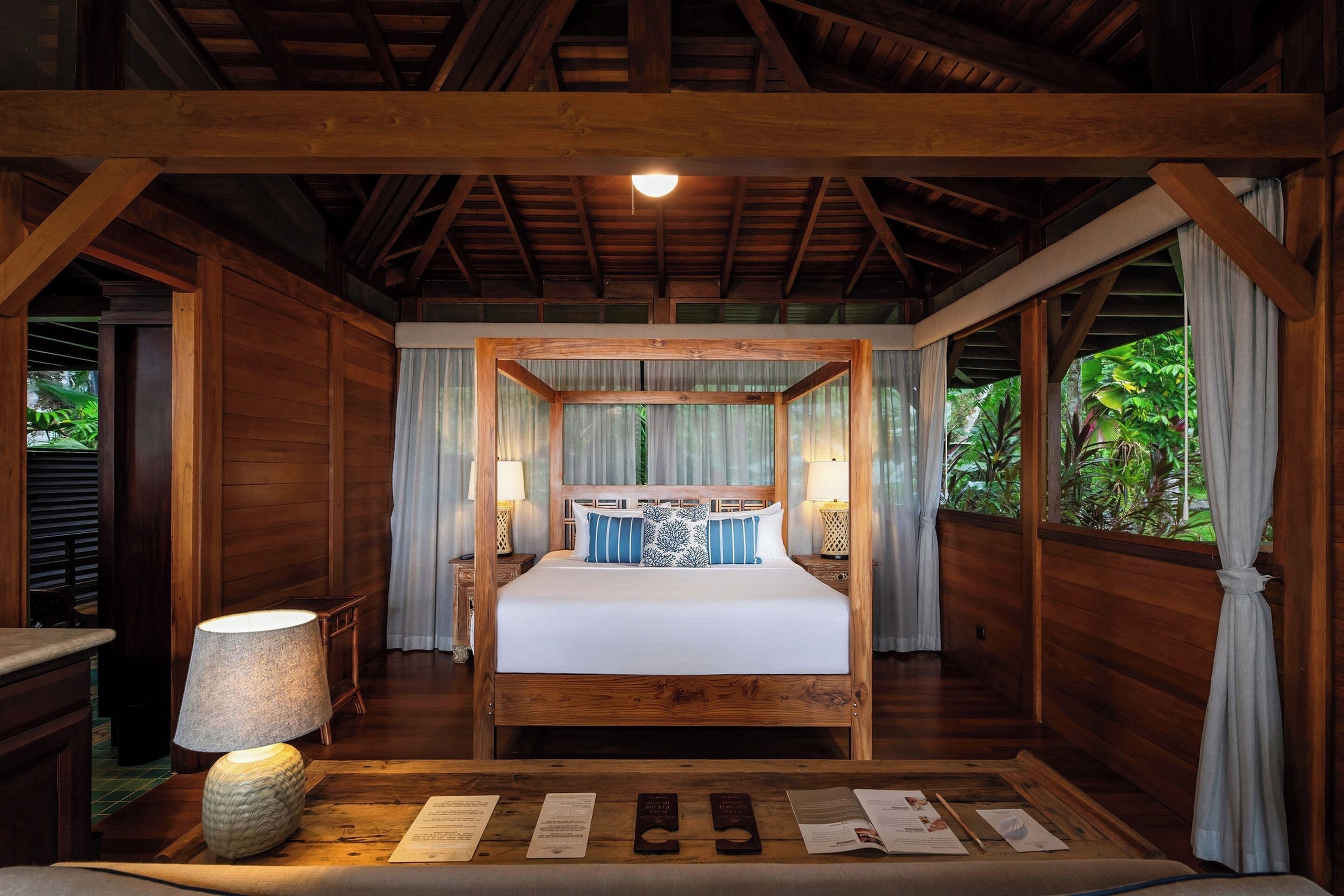 A wooden four-poster bed with white bedding and blue pillows, flanked by two bedside tables with lamps, in a room with wooden walls and a sloped ceiling.