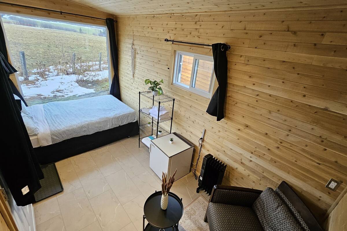 Ein gemütliches Blockhauskabinenzimmer mit einem Bett, einem kleinen Tisch mit einer Vase, einer Heizung und Blick auf eine verschneite Landschaft.