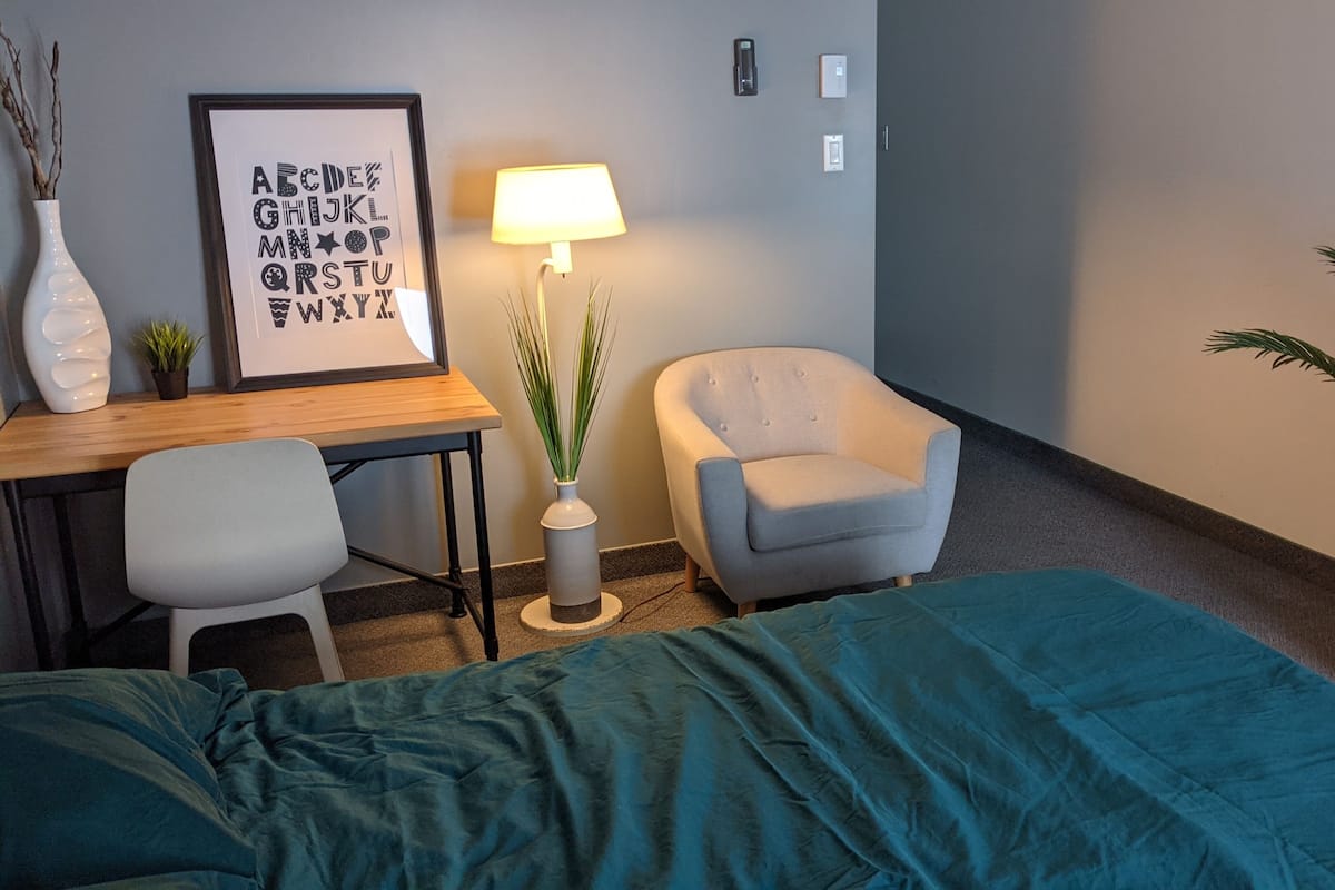 Ein ordentlich eingerichtetes Hotelzimmer mit einem Bett, einem Schreibtisch mit Stuhl, einer Lampe und einem gerahmten Alphabet-Poster an der Wand.
