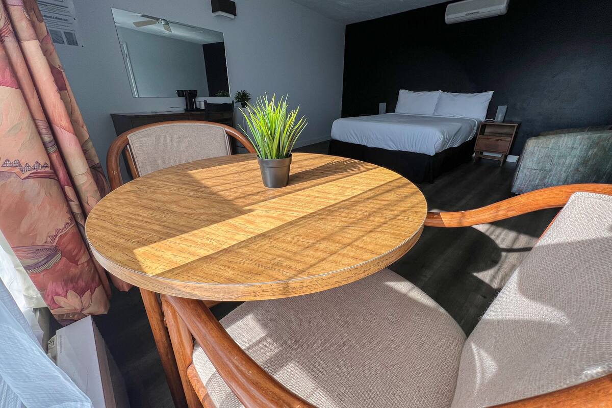 Ein Hotelzimmer mit einem runden Holztisch, einem Blumentopf, einem Bett mit weißer Bettwäsche, einem Nachttisch und einem Spiegel an der Wand.