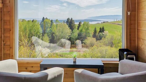 A room with a large window offering a view of a green landscape and distant mountains. There are two white chairs and a small table in front of the window. - Auberge de Nos Aïeux (Les Eboulements)