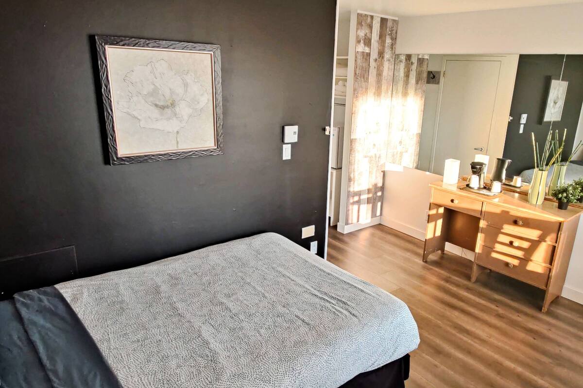 Ein modernes Schlafzimmer mit Bett, Schreibtisch und einem kleinen Tisch. An der Wand befindet sich ein Spiegel und eine Tür zu einem anderen Raum.