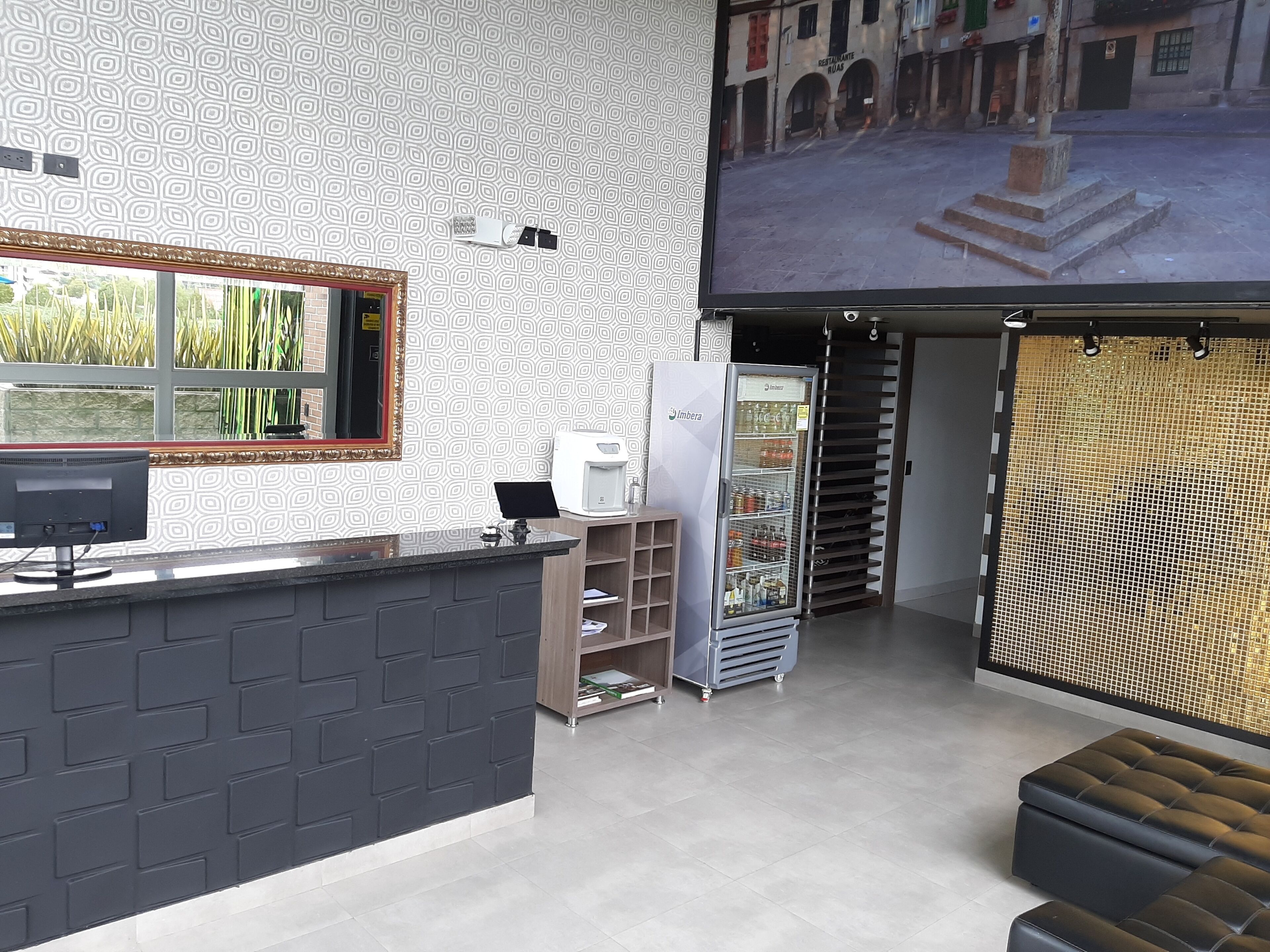 A modern hotel lobby with a reception desk, a large screen displaying a view of the street, a refrigerator, and a seating area.