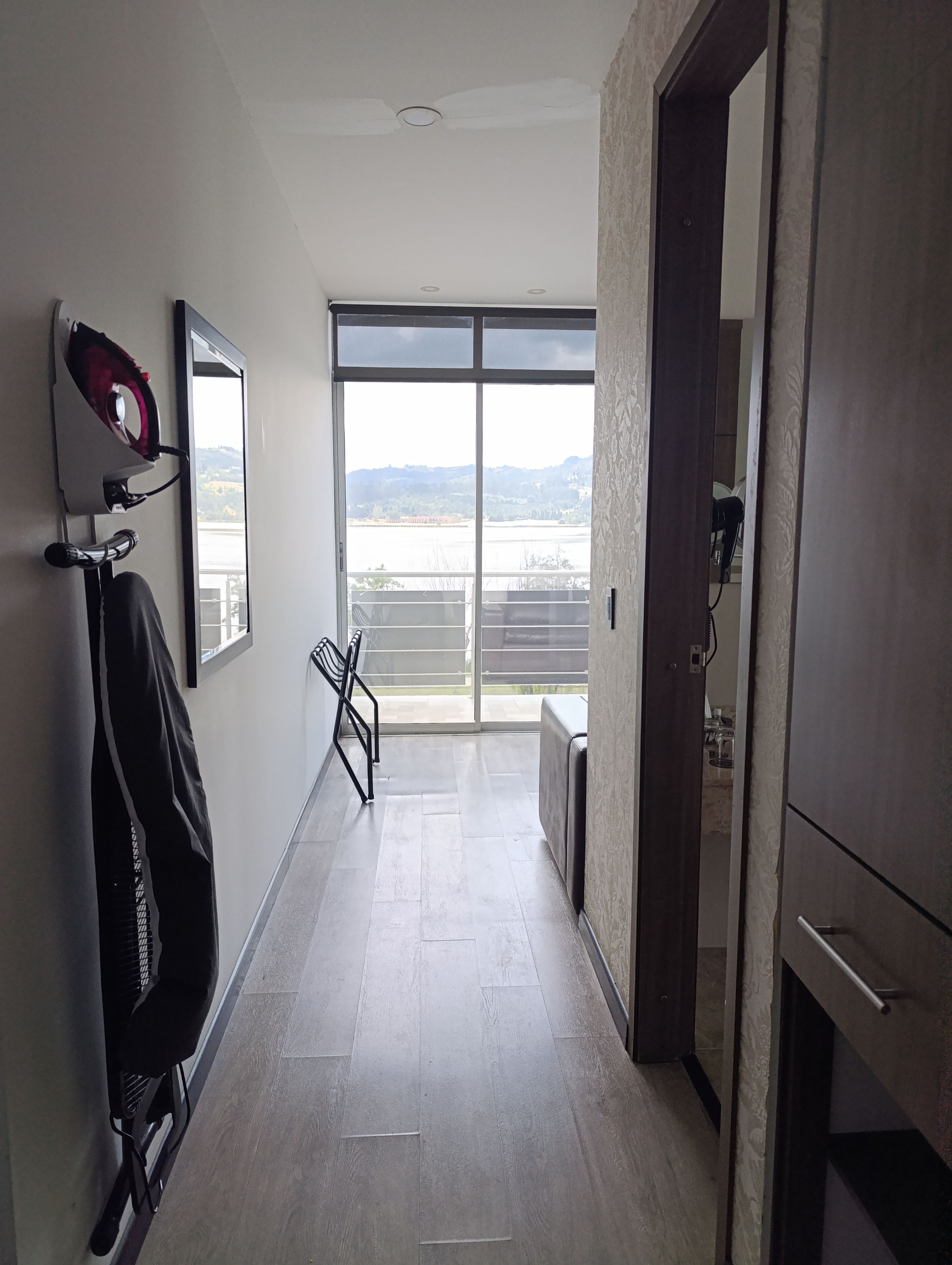 Passadís amb vista a una massa d'aigua i alguns turons, una cadira negra, un penjador i una porta que dona a una habitació.
