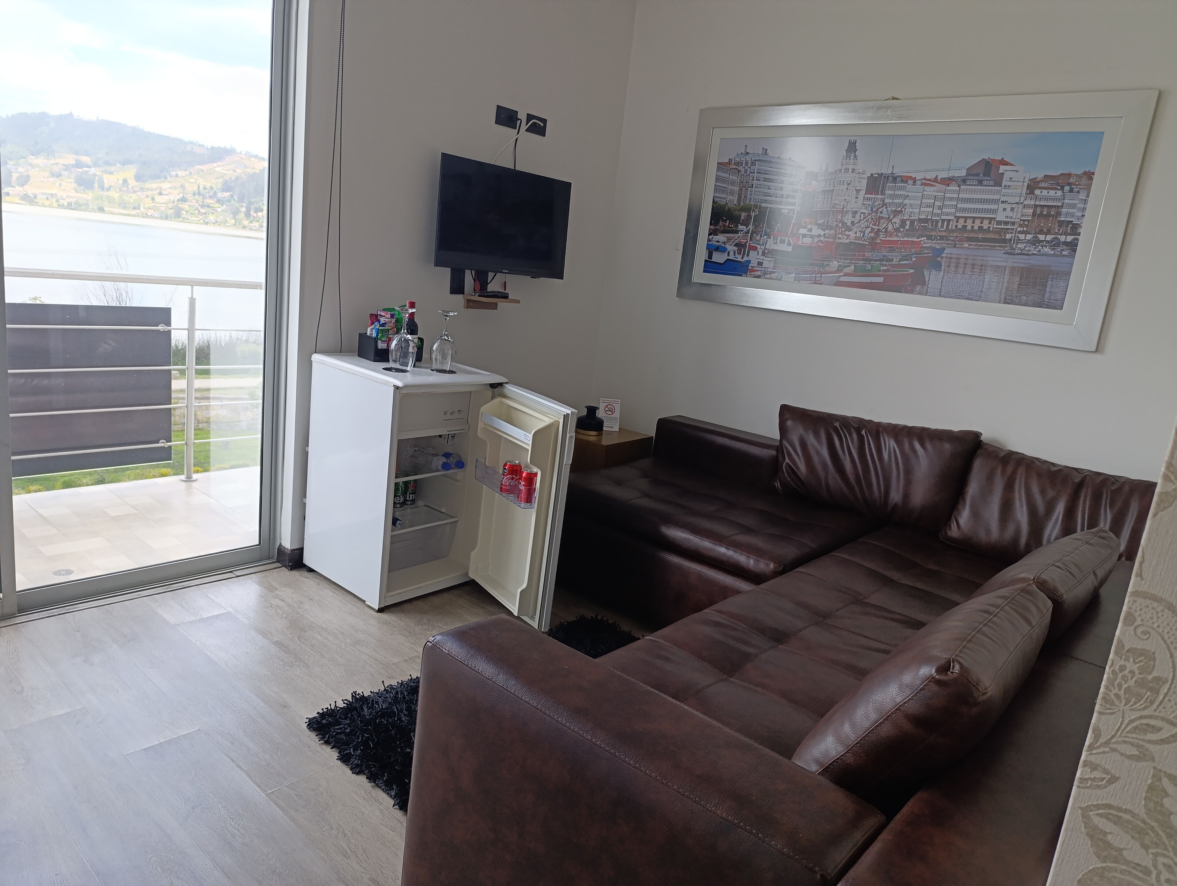 A living room with a brown leather sofa, a white mini-fridge, and a flat-screen TV mounted on the wall.