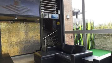 Un interior moderno con un sofá de cuero negro, una escalera y vista a un área exterior verde a través de ventanas amplias.