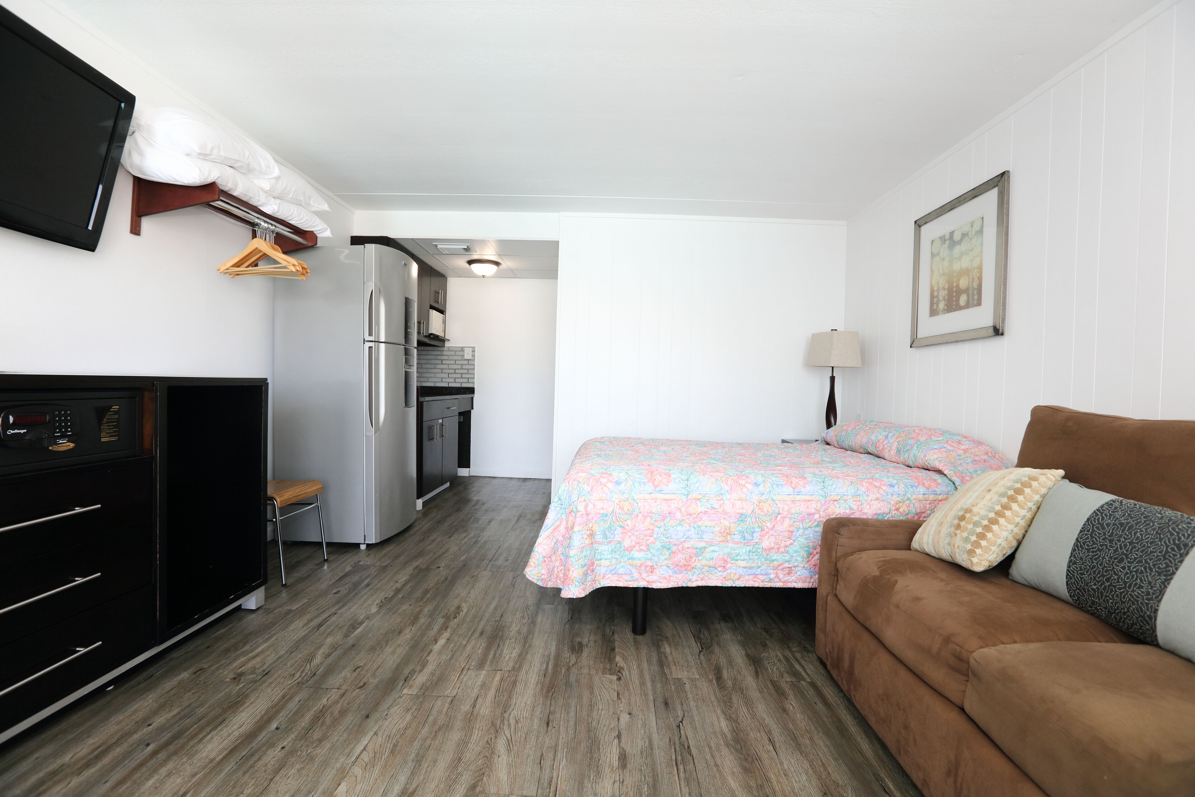 Una habitación de hotel pequeña con kitchenette, una cama con ropa de cama floral, un sofá marrón y un televisor montado.