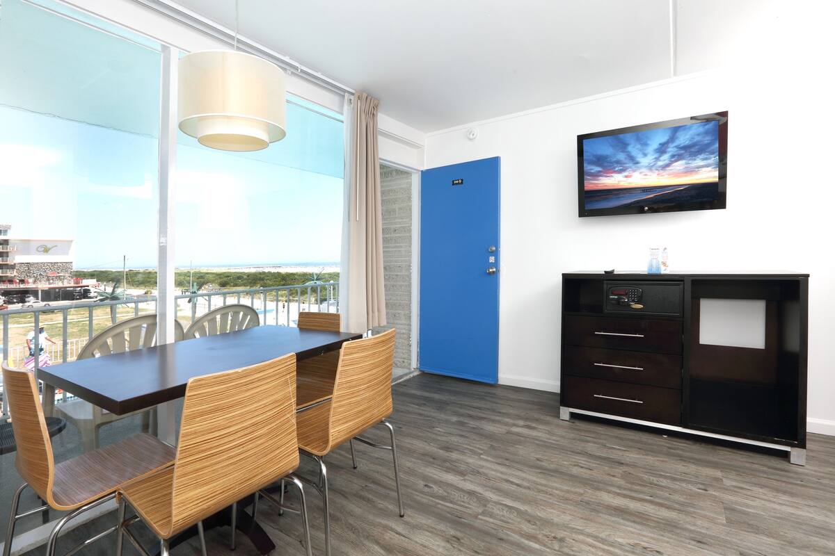 Ein Zimmer mit Esstisch und Stühlen, einer blauen Tür, einem Fernseher an der Wand und Blick ins Freie.