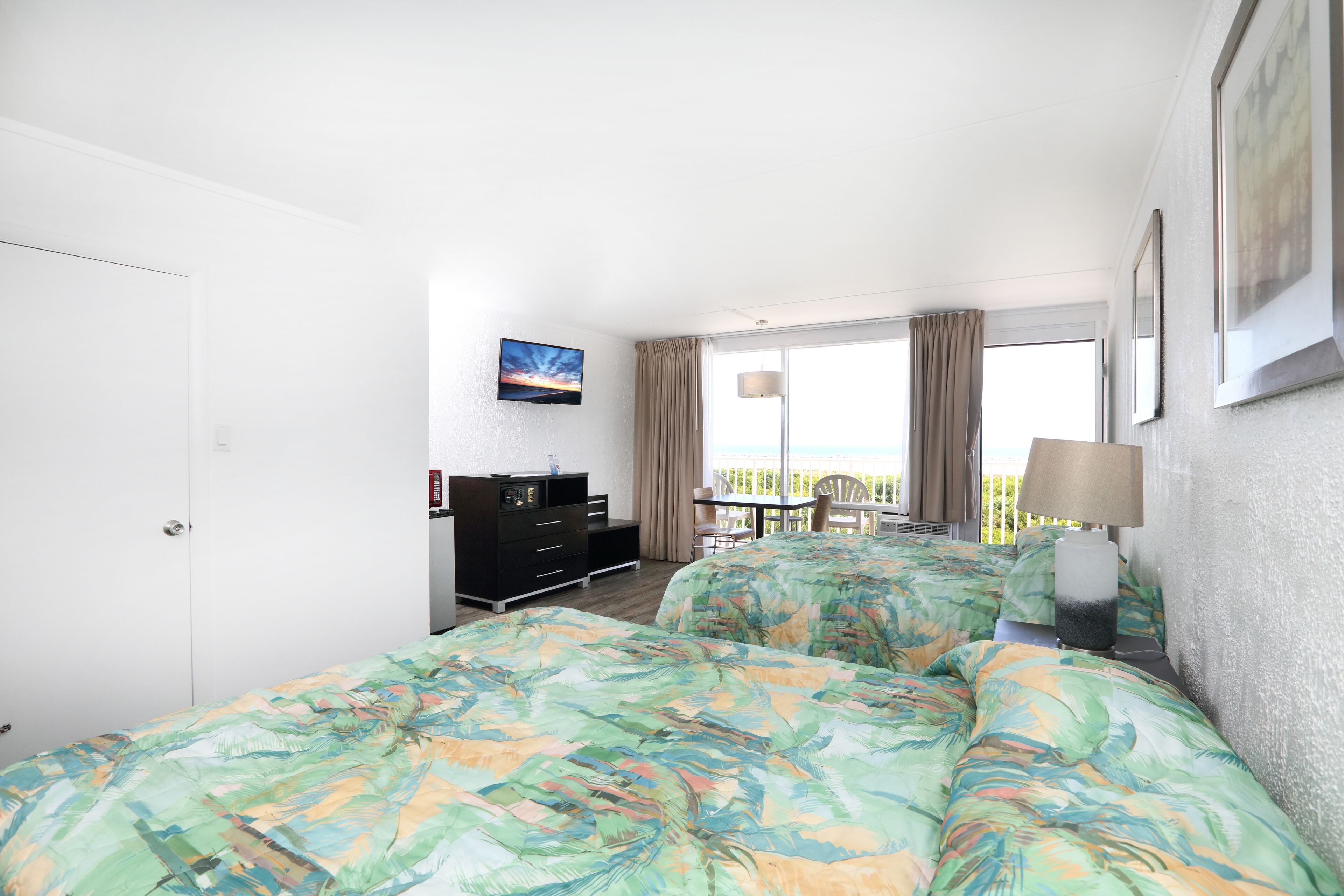 A hotel room with two beds, a desk, a chair, a dresser, and a balcony with a view.