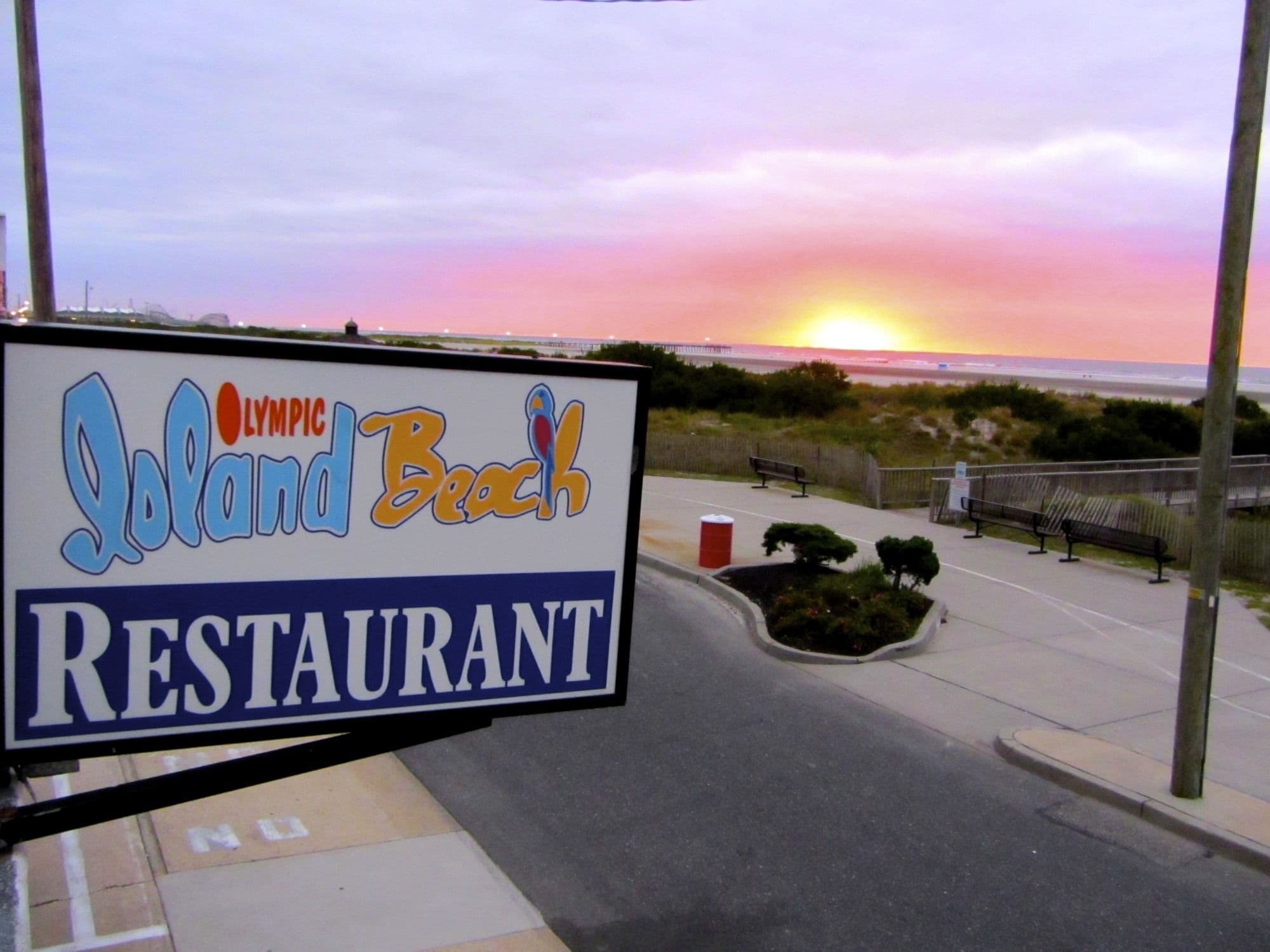 A sign for Olympic Beach Restaurant with a sunset view in the background.