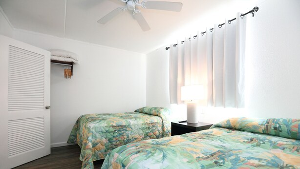 Ein Schlafzimmer mit zwei Betten, einem Deckenventilator, einem Fenster mit Vorhängen und einem Nachttisch mit einer Lampe.