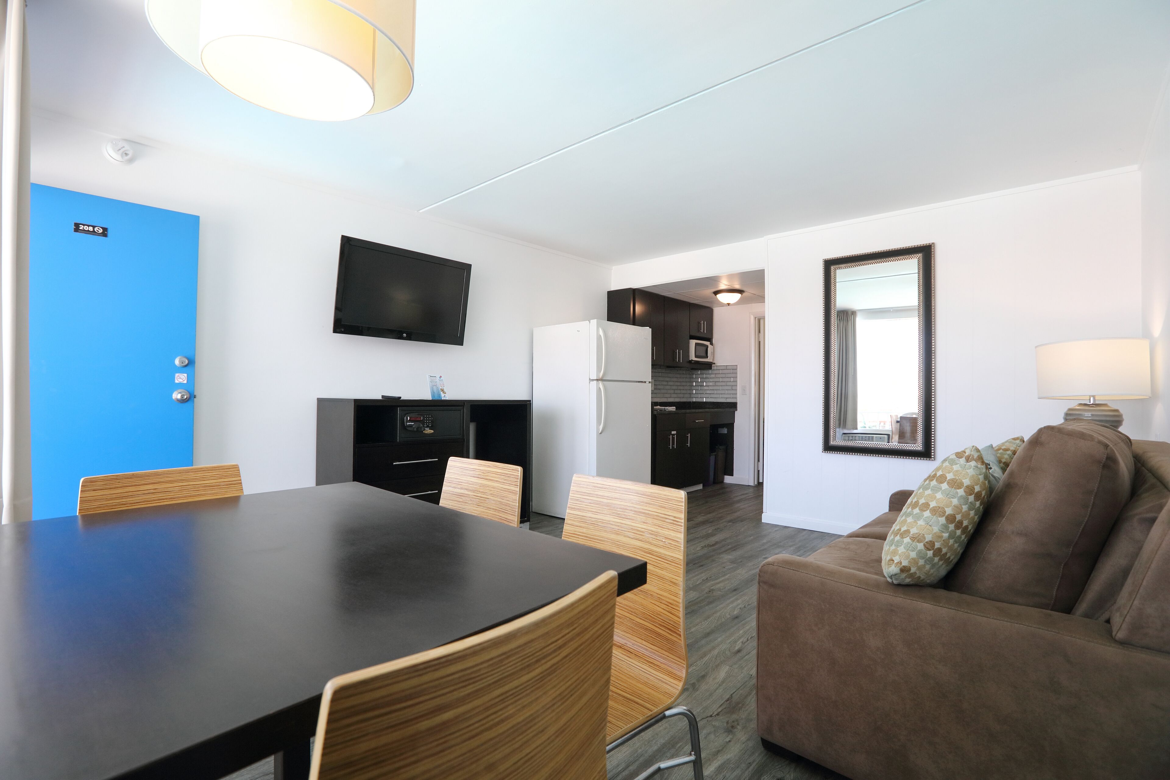 A modern living room with a brown sofa, a dining table with chairs, a kitchen area with a microwave, and a TV mounted on the wall.