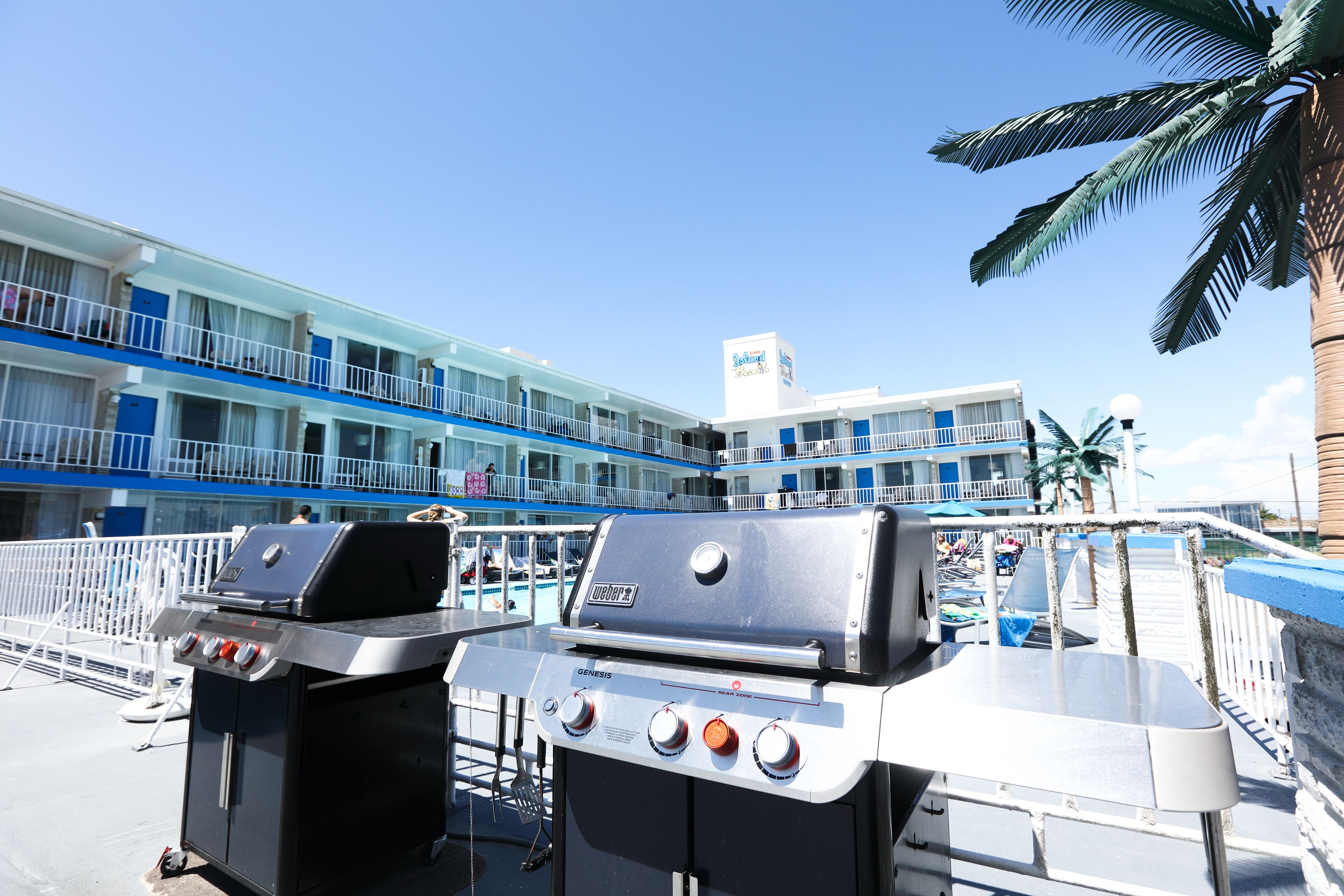 Una parrilla Genesis portátil con pantalla digital, frente a un hotel con decoración azul y blanca, palmeras y un cielo despejado.