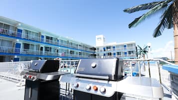 Una parrilla Genesis portátil con pantalla digital, frente a un hotel con decoración azul y blanca, palmeras y un cielo despejado.