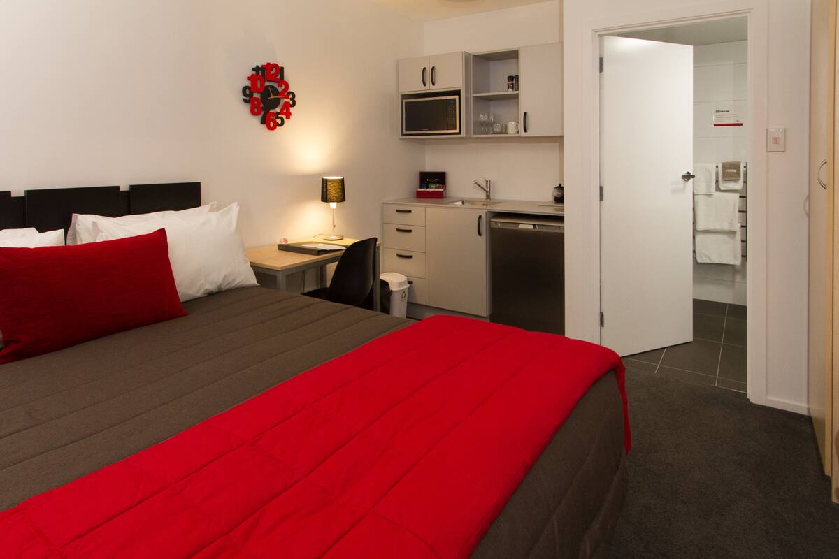 Quarto de hotel com cama, escrivaninha, micro-ondas e uma pequena cozinha.