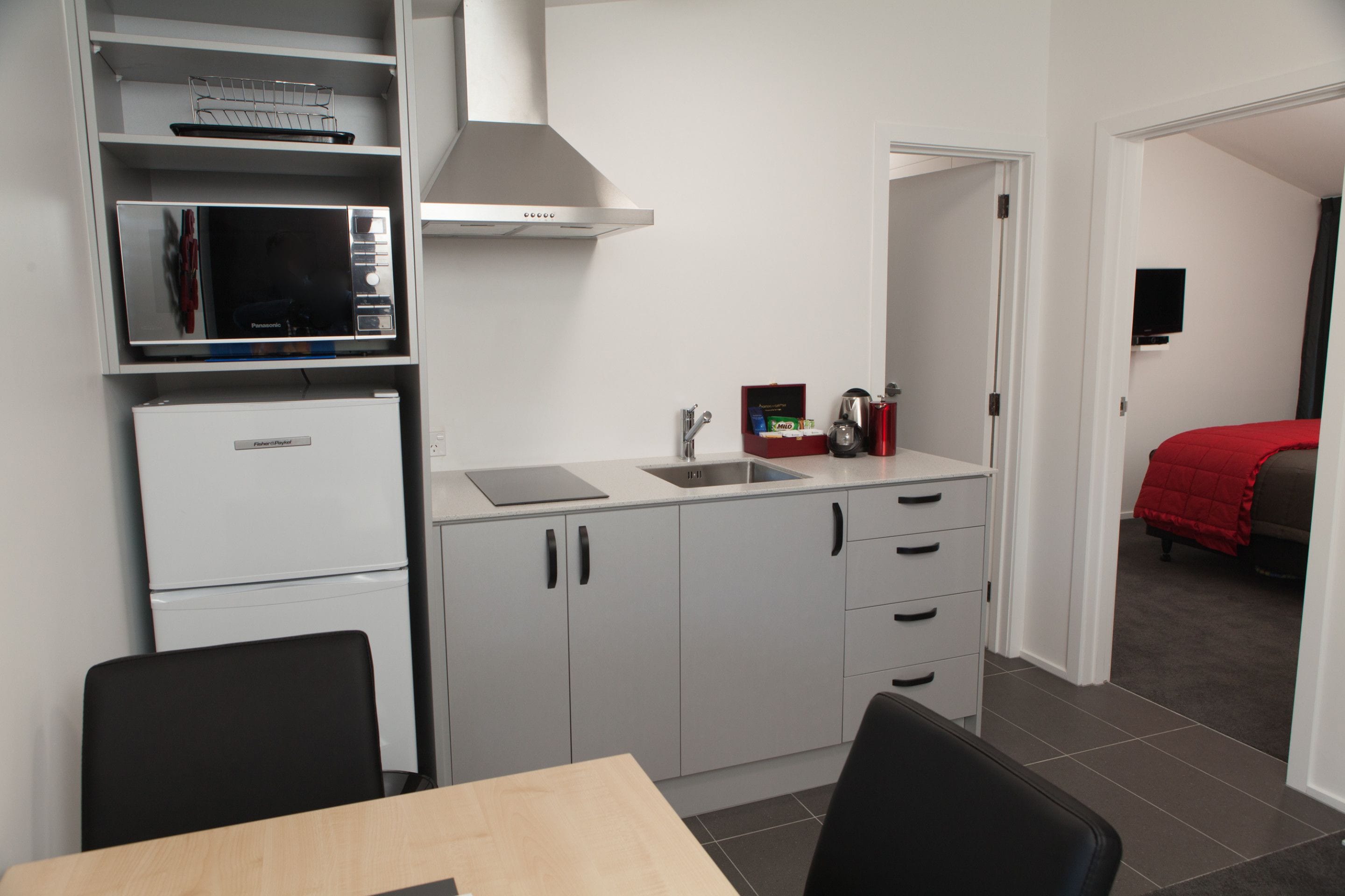 A compact kitchen with a microwave, refrigerator, and sink. There is a dining table with chairs, and a bed with a red blanket in the adjacent room.