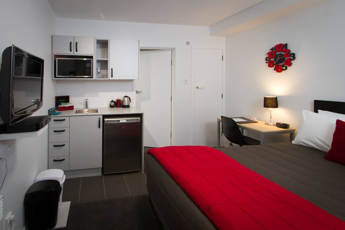 Quarto de hotel pequeno com cozinha compacta, cama com roupa de cama vermelha, escrivaninha com luminária e relógio de parede.