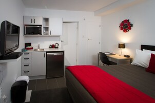 Habitación de hotel compacta con kitchenette, una cama con ropa de cama roja, un escritorio con lámpara y un reloj de pared.