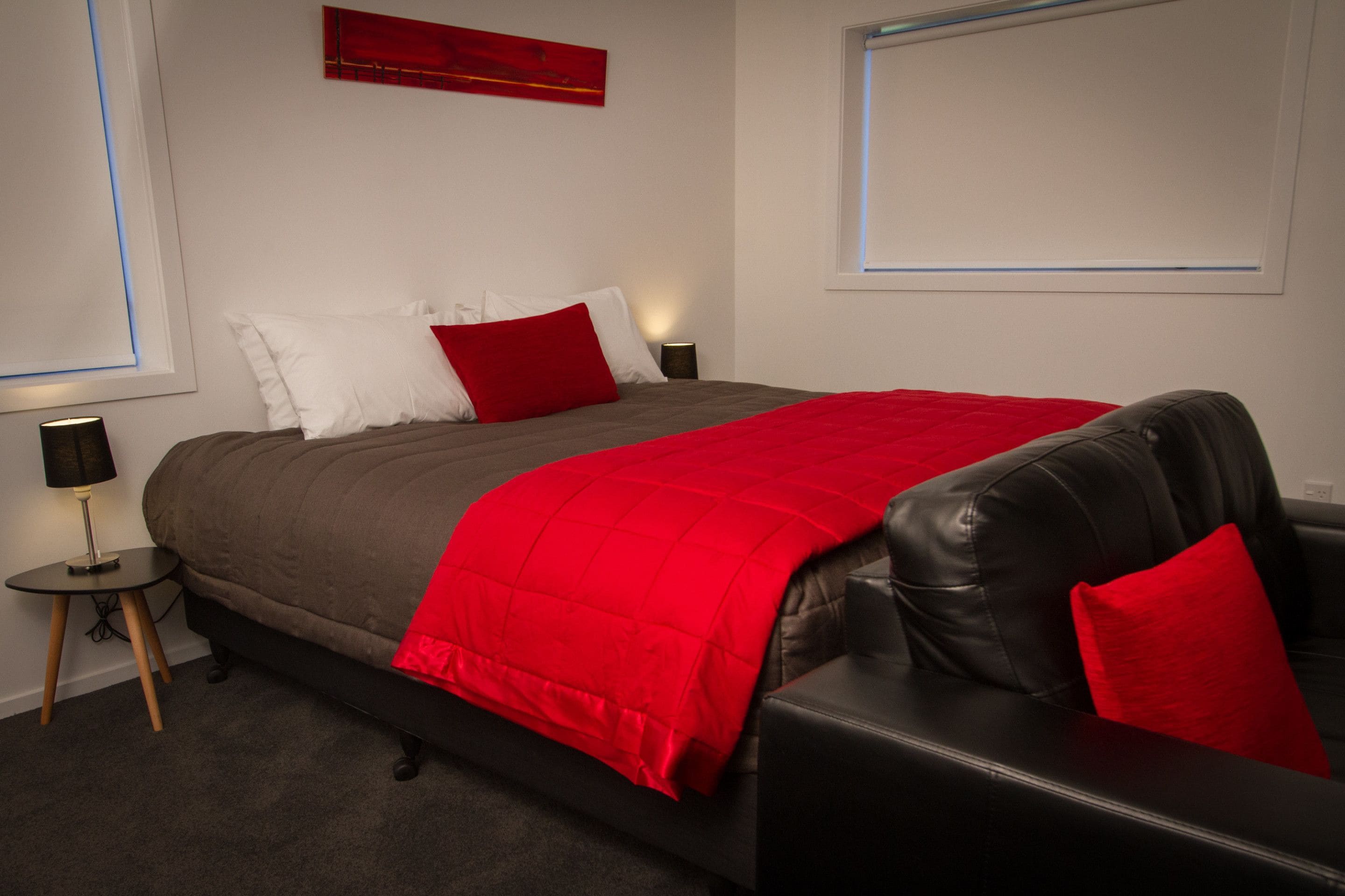 Une chambre à coucher avec un lit, une couvre-lit rouge et marron, un fauteuil en cuir noir, un oreiller rouge, une petite table avec une lampe et une œuvre d’art rouge accrochée au mur.