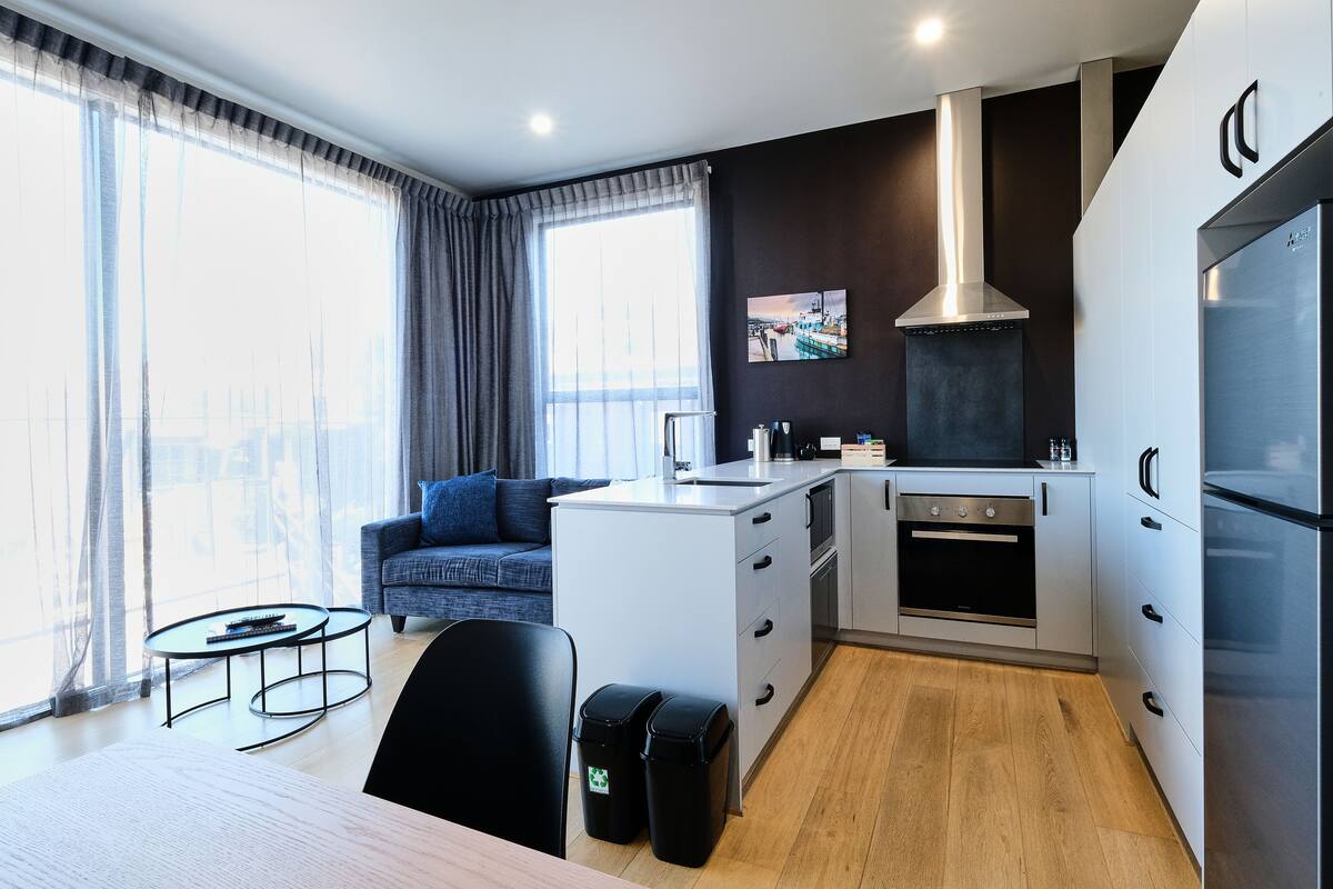 Cozinha moderna com armários brancos, coifa de aço inoxidável e forno embutido. Há uma área de jantar com mesa e cadeiras, e um sofá ao fundo.