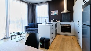 Una cocina moderna con gabinetes blancos, una campana extractora de acero inoxidable y un horno empotrado. Hay un comedor con una mesa y sillas, y un sofá al fondo.