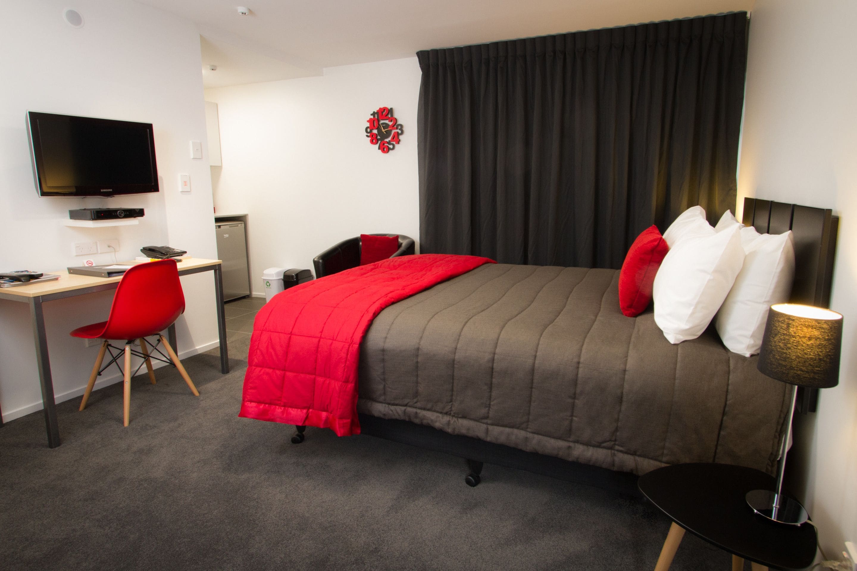 A hotel room with a bed, a desk with a chair, a television, and a clock on the wall.