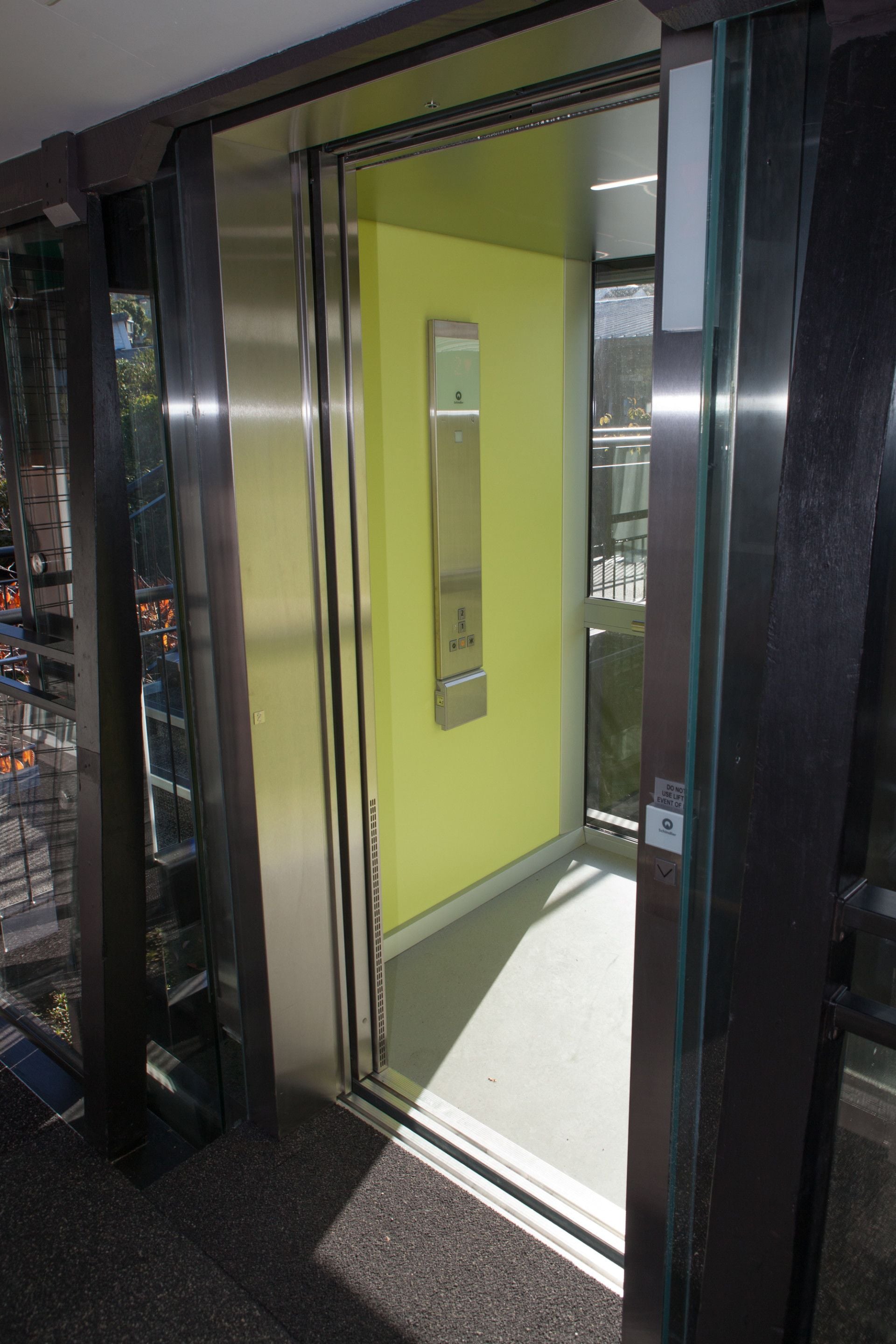 An elevator with a glass door, a green wall, and a metal handrail.