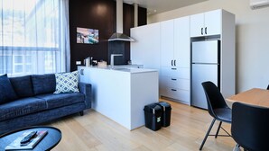 Una cocina moderna con armarios blancos, una estufa integrada y un refrigerador. Hay un comedor con mesa de madera y sillas negras. Un sofá con un cojín estampado se encuentra en la sala de estar, y un ventanal proporciona luz natural.