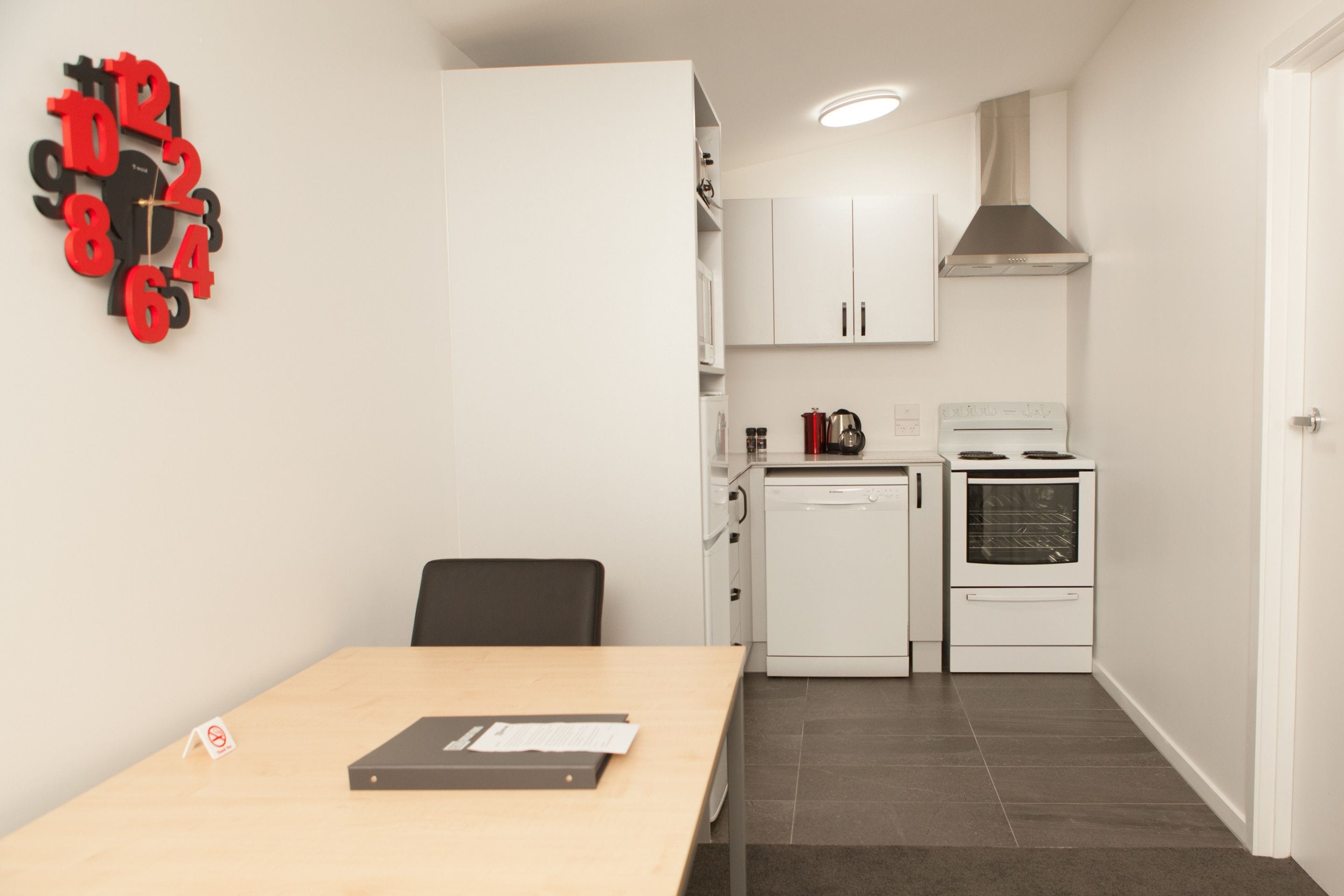 Een kleine keuken met witte kastjes, een fornuis en een magnetron. Er staat een eettafel met een stoel en een wandklok.