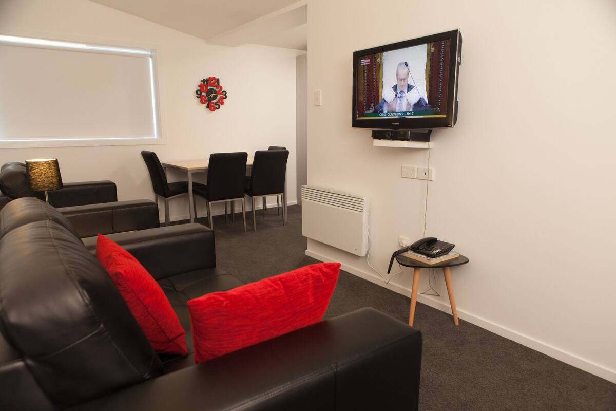 Uma sala de estar moderna com sofá de couro preto, almofadas vermelhas, televisão montada na parede, área de jantar com mesa e cadeiras, e um banquinho de madeira com um telefone.