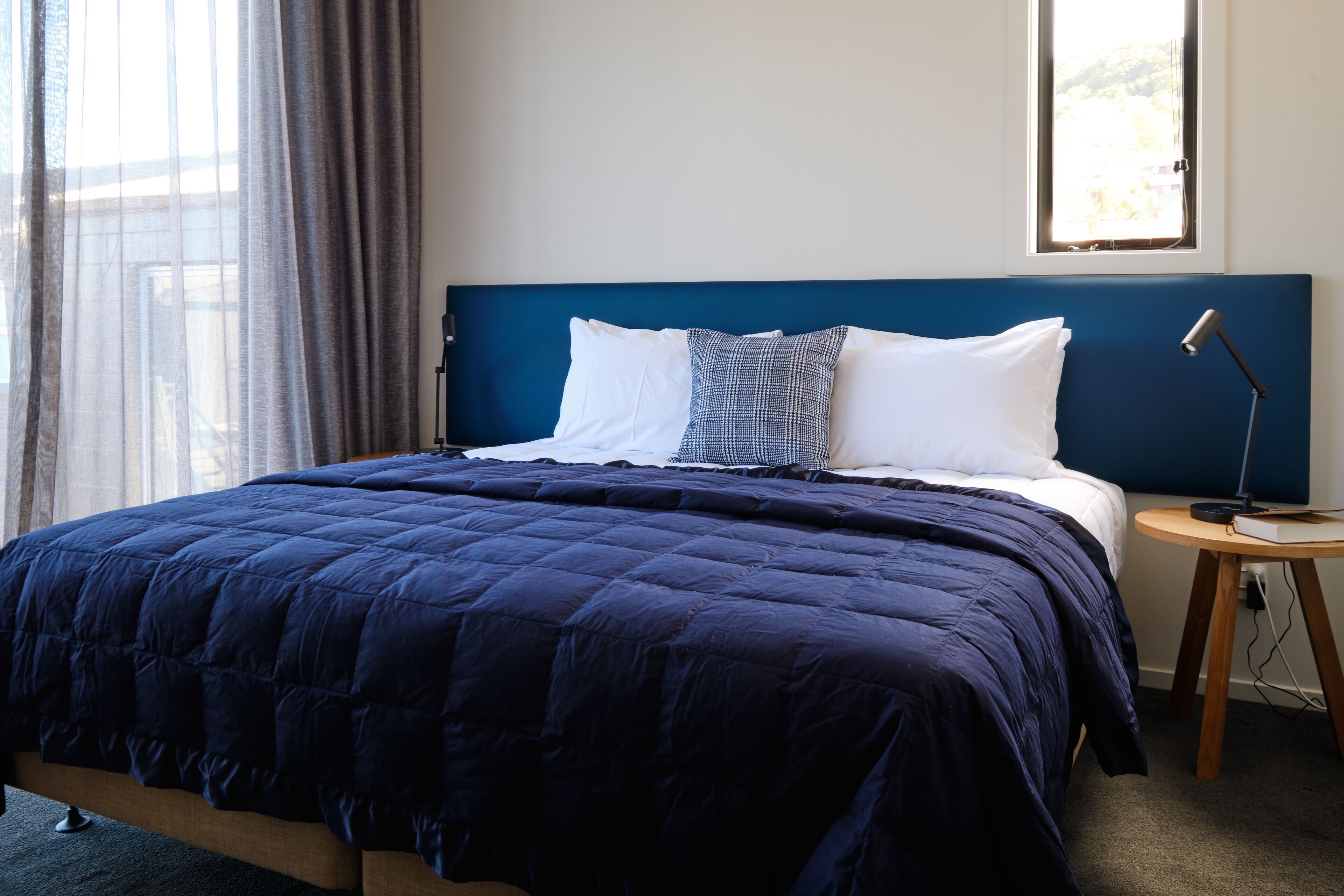 Una cama bien hecha con colcha azul oscuro, un almohadón blanco y un cojín estampado. Se observa una mesita de noche con una lámpara y un libro. En el fondo, se ve una ventana con cortinas.