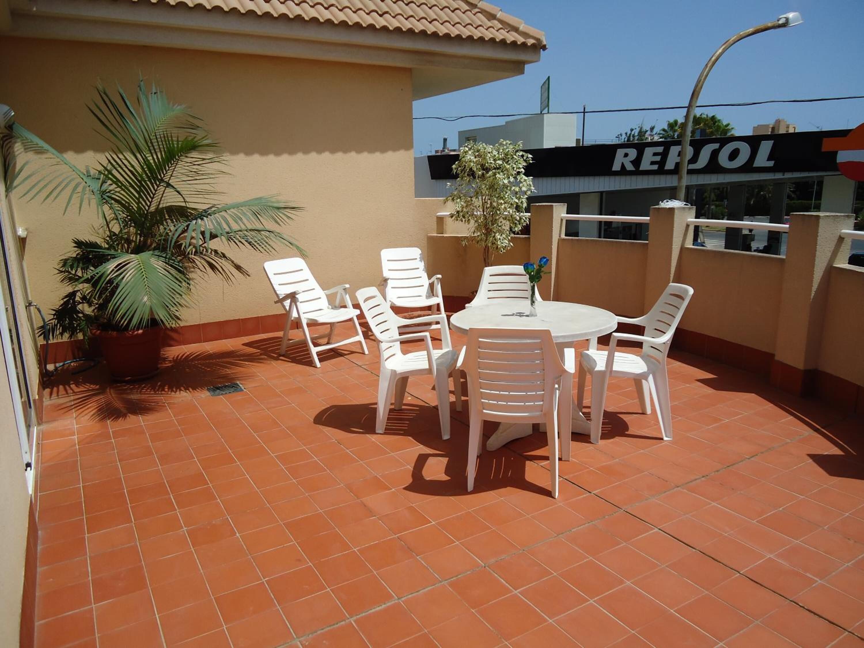 An outdoor patio with a round table and white chairs, a potted plant, and a Repsol sign in the background.