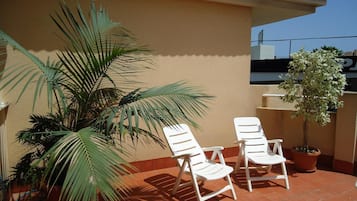 A terrace with two white lounge chairs, a potted palm tree, and a small tree in a pot.