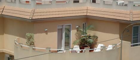 A building with a cafe on the ground floor, a balcony with white chairs, and a palm tree.