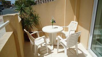 A small outdoor balcony with a round table and four white plastic chairs. There is a potted plant to the left and a glass door to the right.