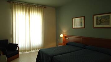 Habitación de hotel con dos camas, una silla, una mesita de noche con lámpara, y dos cuadros en la pared.