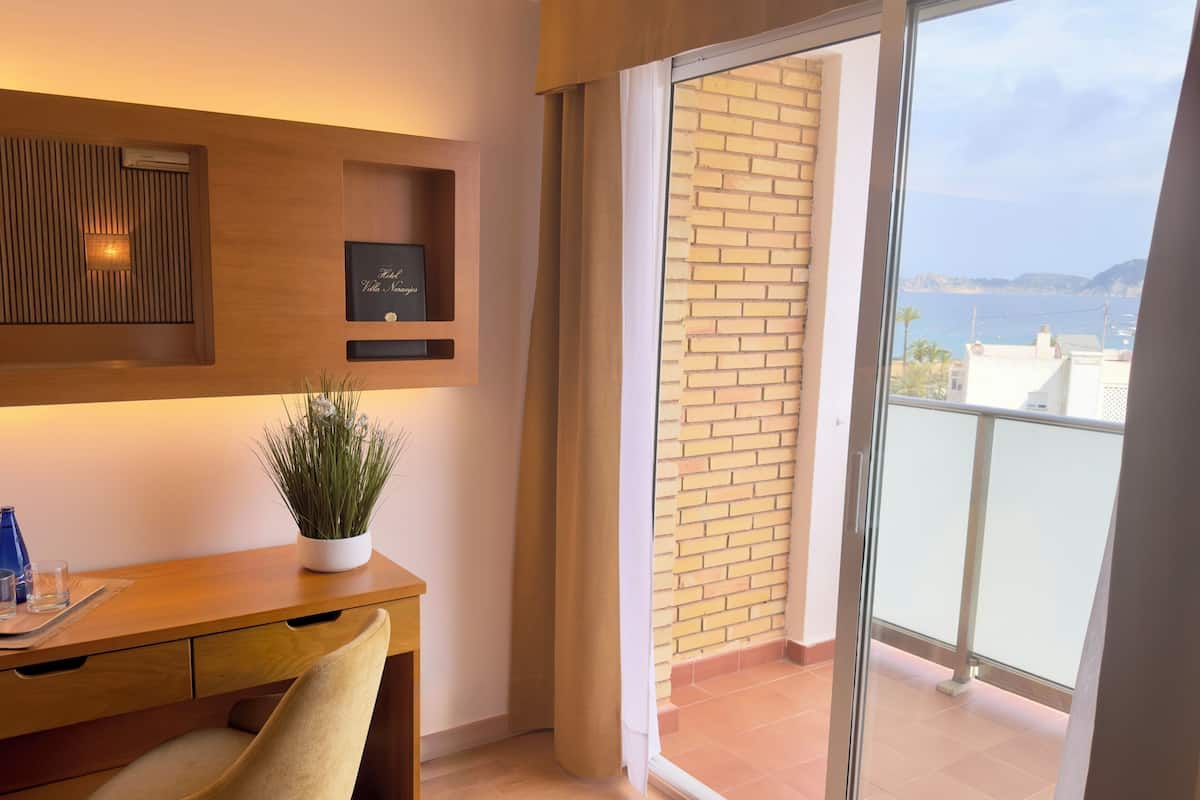 A modern room with a wooden desk, a potted plant, a framed picture, and a view of the sea through a balcony.