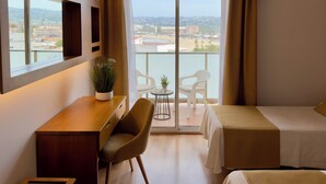 Habitación de hotel con escritorio, dos sillas, una cama, una mesita con planta y un balcón con vista a la ciudad.