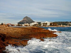 A coastal hotel with a prominent mountain in the background, rocky shoreline, and waves crashing onto the beach. - Hotel Villa Naranjos (Javea)