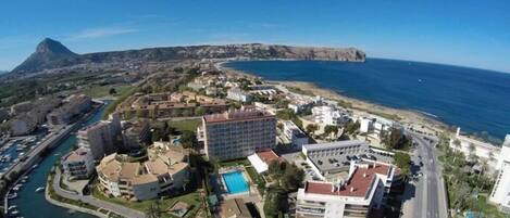 Un complejo turístico frente al mar con un puerto deportivo, un gran edificio de hotel y una montaña al fondo.