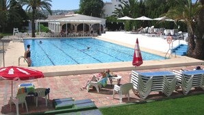 Una piscina con sillas de descanso y sombrillas, rodeada de palmeras y un edificio de fachada blanca.